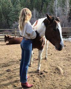 a woman standing next to a brown and white horse on top of a dirt field