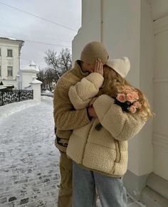 a man and woman hugging in front of a building on a snow covered street with houses in the background