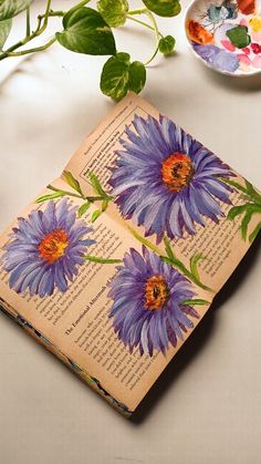 an open book sitting on top of a table next to a plate with flowers painted on it