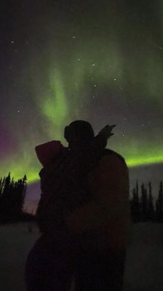 two people are standing in the snow with an aurora light behind them and trees on the other side