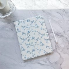 a white and blue flowered notebook on a marble counter top next to a candle