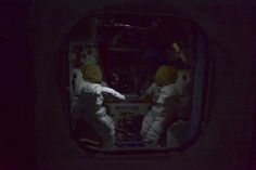 two astronauts in space suits are seen through the window of a space station at night