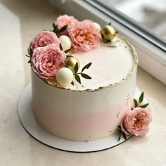 a white cake with pink flowers on top and gold trim around the edges, sitting in front of a window