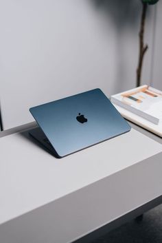 an apple laptop sitting on top of a white table next to a book and plant