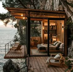 a wooden deck with chairs on it next to the water and a tree covered cliff