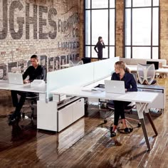 two people sitting at desks in an office with large windows and brick wall behind them