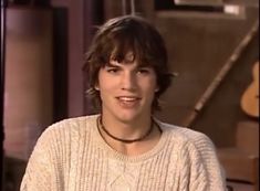 a young man wearing a white sweater sitting in front of a tv screen with his hand on his hip