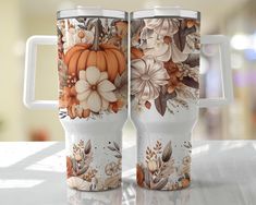 two coffee mugs decorated with flowers and pumpkins on a white countertop in front of a blurry background