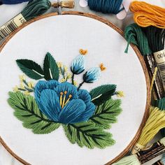 a blue flower with green leaves on it in a wooden hoop next to yarn and thread