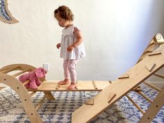 a toddler girl dressed in white top and pink leggins stands in the center of an indoor wooden playground containing a climbing arch, climbing triangle, and a bridge connecting the two. She is playing with a bright pink stuffed puppy. Toddler Playroom Ideas, Toddler Climbing Wall, Climbing Toys For Toddlers, Toddler Climbing Toys, Toddler Climbers, Climbing Arch, Toddler Climbing, Montessori Bedroom, Pikler Triangle