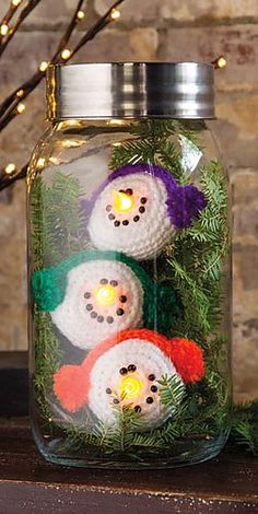 three crocheted snowmen in a glass jar with lights on the side and text below