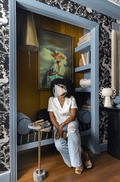 a woman sitting in a room with a painting on the wall and shelves behind her