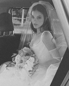 a woman sitting in the back seat of a car wearing a wedding dress and holding a bouquet of flowers