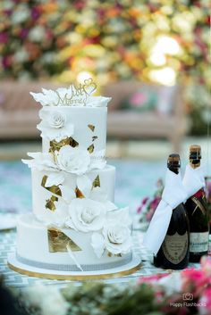 a wedding cake with white flowers and gold accents sits next to a bottle of champagne