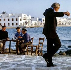 a man in a suit and hat is dancing by the water with other men sitting at tables