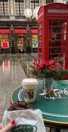 a woman holding a cup of coffee in front of a red phone booth with flowers