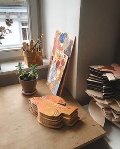 a wooden table topped with lots of paintings and papers next to a potted plant