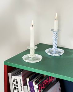 two white candles are sitting on a green book shelf next to a stack of books