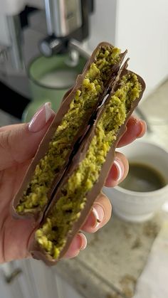 a person holding up a chocolate and pistachio sandwich in front of a cup of coffee