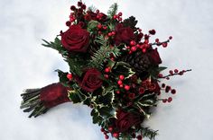 a bridal bouquet with red roses and greenery on the snow covered ground next to pine cones