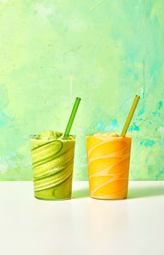 two drinks with straws sitting next to each other on a white counter top in front of a green wall