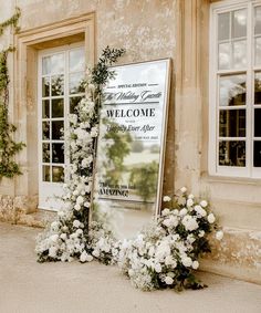 a welcome sign with flowers and greenery in front of two windows on the side of a building