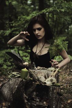 a woman in black dress sitting on a tree stump with plants and candles around her