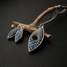 two pendants are hanging from a branch on a black surface, one is blue and the other is silver