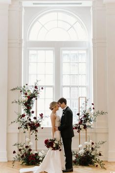 a bride and groom standing next to each other in front of a window with red flowers