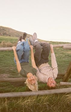 two people laying on the ground in front of a wooden fence with grass and hills behind them