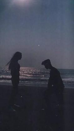 two people are standing on the beach at night, one is looking at the water