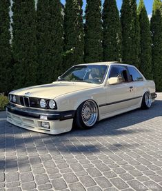 a white car parked on top of a brick road next to tall green trees and bushes