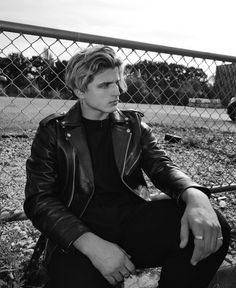 a young man sitting on the ground next to a chain link fence wearing a leather jacket