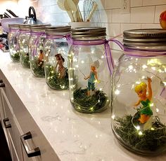 several glass jars with fairy figurines in them on a kitchen counter, one is filled with grass and the other has lights