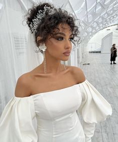 a woman with curly hair wearing a white dress and pearls on her head, standing in front of a wall