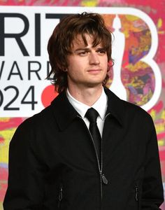 a young man in a black jacket and tie standing next to a colorful wall with the brit awards logo on it