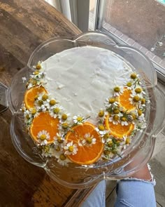 a cake decorated with oranges and daisies on a glass plate in front of a window