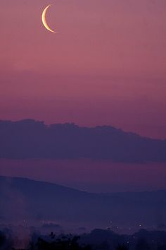 the moon is setting in the purple sky above some hills and trees, with mountains in the background