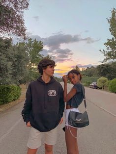 two young people standing on the side of a road with trees and bushes in the background