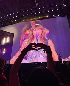 a person holding up a heart shaped hand in front of a large screen with a woman on it