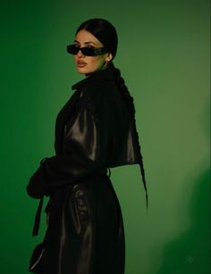 a woman wearing sunglasses and a black leather coat is posing for the camera in front of a green background