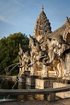 an elephant fountain in front of a building with statues on it's sides and trees behind it