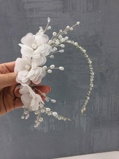 a hand holding a white flower and pearled headpiece in front of a gray wall