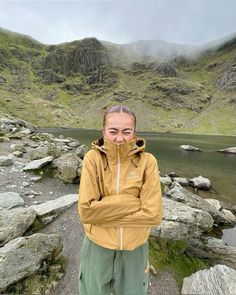 a woman standing on the side of a mountain next to a lake and mountains in the background