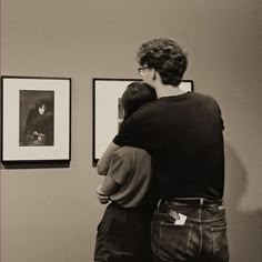 two people are looking at pictures on the wall in an art gallery, one is hugging the other's back