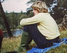 a young man sitting on the grass next to a river with his arms resting on his knees