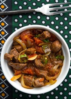 a white bowl filled with meat and onions on top of a green table cloth next to silverware