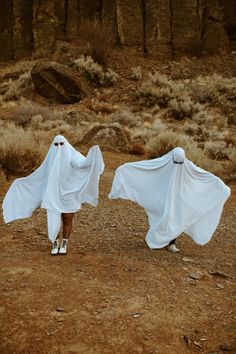 two people in white ghost costumes walking across a dirt field with their arms spread out