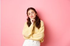 a woman in a yellow sweater is leaning against a pink wall with her hand on her chin