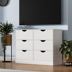 an entertainment center with white drawers and plants in front of a flat screen tv mounted on the wall